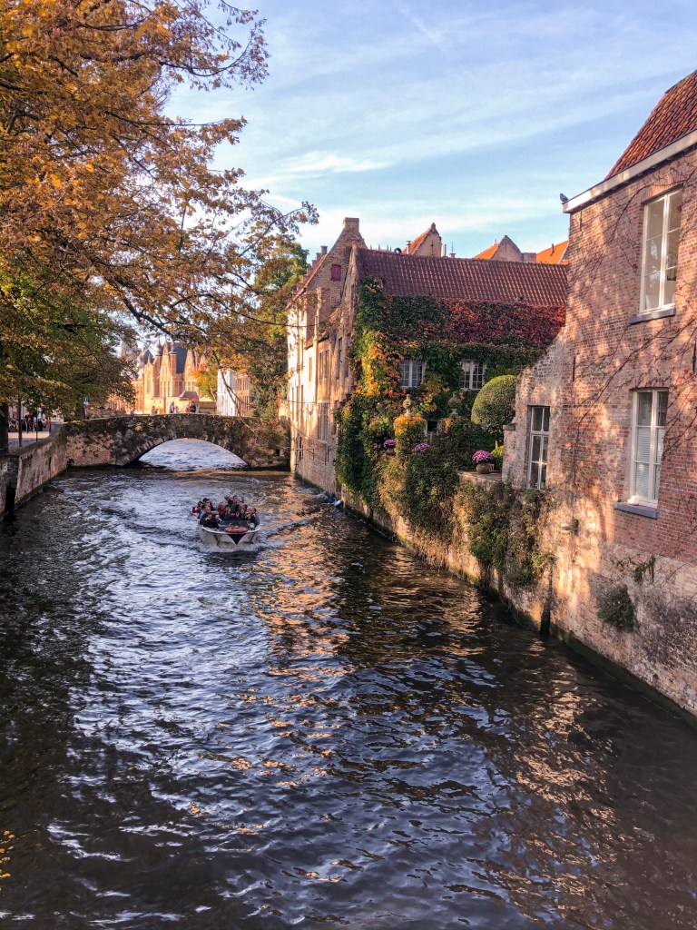 A canal with a bridge crossing it, and an orange tree shading over it. Across the canal there is a garden full of greens and reds of autumn.